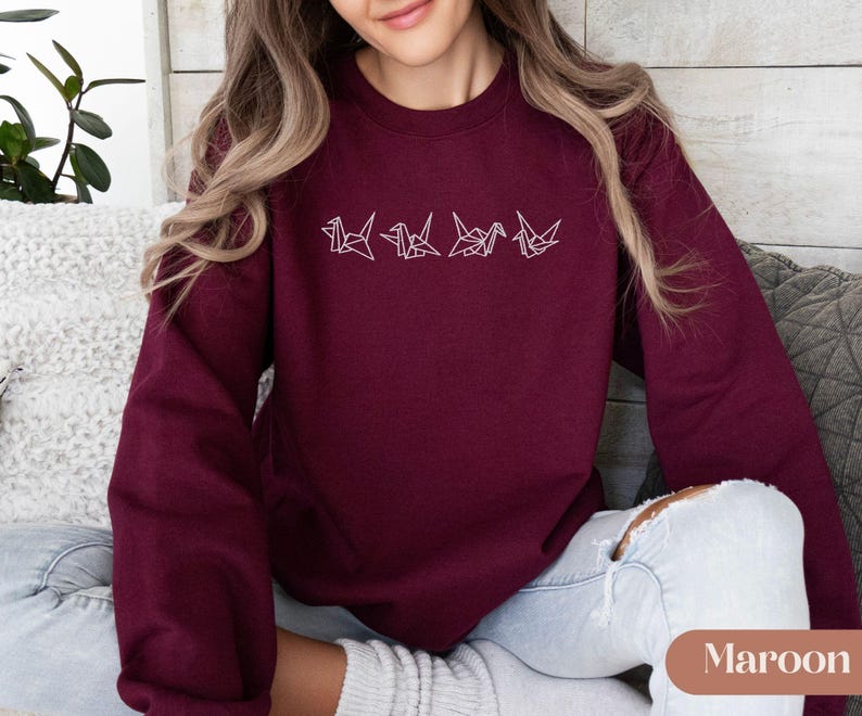 May include: Maroon-colored sweatshirt featuring a minimalist design of four origami cranes in white thread. The sweatshirt has a crew neck and long sleeves. The word "Maroon" is displayed in a small rectangular label at the bottom right.