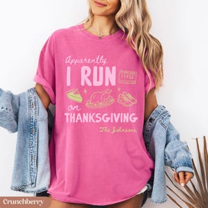K&ouml;nnte beinhalten: Ein leuchtend pinkfarbenes Kurzarm-T-Shirt mit dem Text "Apparently I RUN on THANKSGIVING" in Wei&szlig; und Hellrosa. Die Grafik zeigt ein St&uuml;ck Kuchen, einen gebratenen Truthahn, eine Marathon-Startnummer 15432 und Laufschuhe in Gelb.