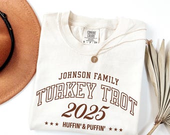 May include: Cream-colored crew neck t-shirt with the text "JOHNSON FAMILY TURKEY TROT 2025 HUFFIN' & PUFFIN'" in brown lettering. A gold necklace with a pendant is draped on the shirt. A brown hat and dried plant are in the background.