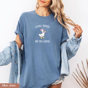 Puede incluir: Camiseta azul vaquero con el texto "LEGAL GOOSE ON THE LOOSE" sobre un ganso de dibujos animados con un lazo rosa y una bebida. La camiseta tiene mangas enrolladas y un ajuste relajado. Una chaqueta vaquera est&aacute; sobre los hombros.