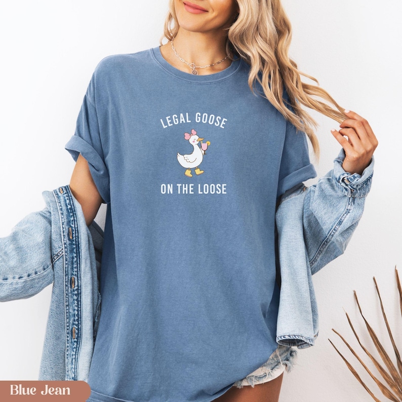 Puede incluir: Camiseta azul vaquero con el texto "LEGAL GOOSE ON THE LOOSE" sobre un ganso de dibujos animados con un lazo rosa y una bebida. La camiseta tiene mangas enrolladas y un ajuste relajado. Una chaqueta vaquera est&aacute; sobre los hombros.