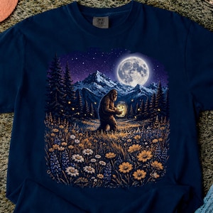 Könnte beinhalten: Ein dunkelblaues T-Shirt mit einem Grafikdruck eines Bigfoot-Wesens, das nachts auf einem Feld mit Wildblumen steht. Die Szene zeigt einen Vollmond, schneebedeckte Berge, Kiefern und Glühwürmchen, die in einem Glas leuchten.