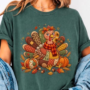 May include: A dark green t-shirt featuring a graphic of a turkey wearing a plaid scarf and a floral crown. The turkey has patchwork-style feathers in various patterns, including leopard print and plaid, surrounded by pumpkins, acorns, and leaves.