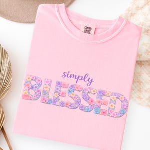Puede incluir: Camiseta rosa claro con las palabras "simply BLESSED" en un diseño floral. La camiseta tiene cuello redondo y la marca "COMFORT COLORS" es visible en la etiqueta.