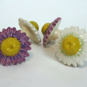 May include: Three ceramic flower-shaped drawer pulls with yellow centres and white, purple, and green petals.