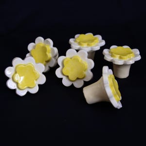 May include: Six ceramic bottle stoppers with a daisy design. Each stopper has a white, petal-shaped rim and a yellow center. The stoppers are displayed against a black backdrop, highlighting their floral design.