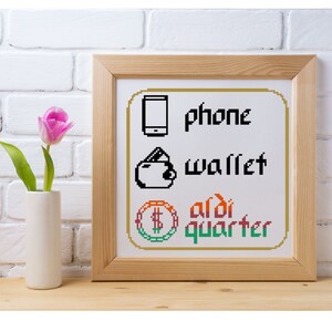 Puede incluir: Una obra de arte en punto de cruz enmarcada con las palabras "phone", "wallet" y "aldi quarter". La obra presenta imágenes pixeladas de un teléfono, una cartera y una moneda con un signo de dólar. A la izquierda hay un tulipán rosa en un jarrón blanco.