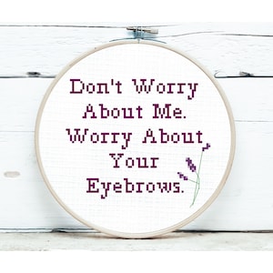 May include: A cross-stitch embroidery hoop with the text "Don't Worry About Me. Worry About Your Eyebrows." in purple. The design includes a small lavender sprig. The hoop is set against a white wooden background.