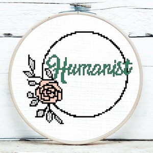 Pode incluir: Um bastidor de bordado em ponto cruz com a palavra "Humanist" em verde, dentro de um círculo preto pixelizado. Uma rosa e folhas pixelizadas rosa e pretas adornam o lado esquerdo. O bastidor está sobre um fundo de madeira branca.