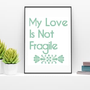 Puede incluir: Una impresión enmarcada con el texto "My Love Is Not Fragile" en letras verdes. Un diseño decorativo se encuentra debajo del texto.