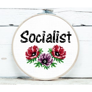Op de afbeelding: Een witte borduurring met het woord "Socialist" in zwarte tekst en een bloemmotief van rode en paarse bloemen met groene bladeren.