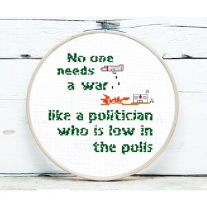 May include: A cross-stitch hoop featuring the text "No one needs a war... like a politician who is low in the polls." The design includes pixelated images of a missile, fire, and a hospital, in green and orange.