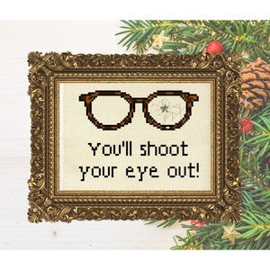 May include: Cross-stitch artwork in a gold ornate frame. The artwork features pixelated brown glasses with a cracked lens and the text "You'll shoot your eye out!" on a cream background. Festive holiday decor surrounds the frame.