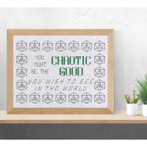 May include: A framed cross-stitch artwork with a light wood frame. The design features geometric shapes with numbers and the words "CHAOTIC GOOD" in green, along with the quote "YOU MUST BE THE GOOD YOU WISH TO SEE IN THE WORLD."