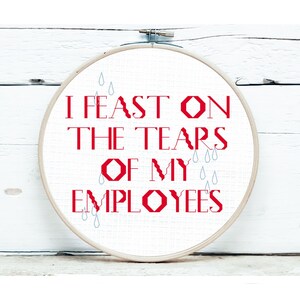 May include: A cross-stitch embroidery hoop with the text "I FEAST ON THE TEARS OF MY EMPLOYEES" in red. The design includes blue teardrop accents. The hoop is set against a white wooden background.