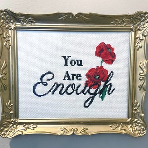 May include: A gold-coloured frame with a white background and black embroidery that reads "You Are Enough". Two red poppy flowers are embroidered on the right side of the frame.