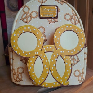 May include: A beige backpack with a Mickey Mouse shaped design. The backpack is covered in a pattern of pretzel shapes and white dots. The backpack has a brown leather tag with the text "Disney Parks" and "Pretzel" on it.