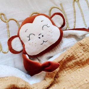 Op de afbeelding: Een pluche aap speelgoed met een bruine kop en oren, een wit gezicht en een glimlachende uitdrukking. De aap is bevestigd aan een zachte, beige en witte deken. De deken heeft een bloemenpatroon en is opgevouwen.