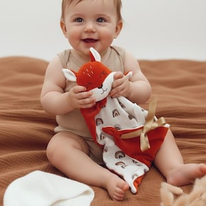 Peut inclure: Un bébé tient une doudou renard rouge et blanche avec un motif arc-en-ciel. Le bébé est assis sur une couverture marron. Un bonnet blanc et des fleurs séchées sont également sur la couverture.