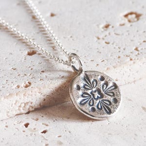 May include: A silver necklace featuring a small, round pendant with a stamped floral pattern. The pendant has a textured, oxidised finish and is suspended from a delicate silver chain, showcasing a symmetrical design with four petals.