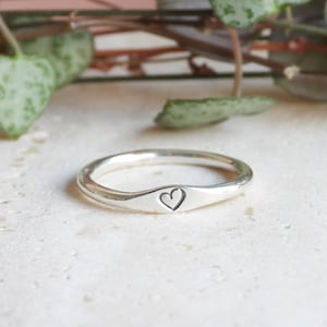 May include: A silver ring featuring a heart motif. The ring has a minimalist design with a polished surface. The heart is engraved on a raised section of the band. The ring is set against a neutral background with green foliage.