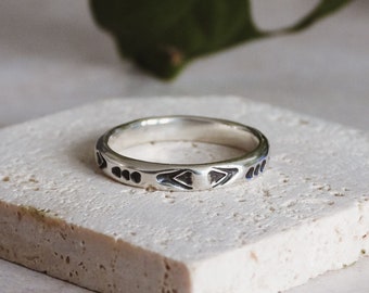 May include: A silver ring with a geometric design. The ring is on a white textured surface.