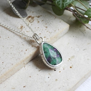 May include: A silver necklace with a teardrop-shaped pendant. The pendant features a green and purple gemstone set in a silver bezel. The necklace is displayed on a textured, light-colored surface with greenery in the background.