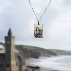 May include: A silver pendant necklace featuring a rectangular charm with a detailed relief of a building. The charm hangs from a delicate chain. The background is blurred, showing a coastal scene with a church.