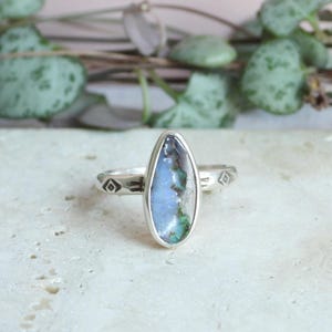 May include: A silver ring with an oval-shaped stone. The stone has a light blue and green color scheme. The ring band has a geometric design on each side of the stone.