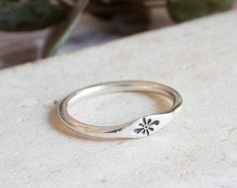 May include: A silver ring with a floral design. The ring is thin and has a slightly curved band. The floral design is etched into the ring's surface. The ring is displayed on a light-colored surface.