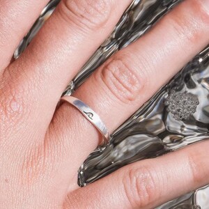 May include: A silver ring with a wave design on a person's finger. The ring is worn on the ring finger of the left hand. The hand is resting on a surface that appears to be water.
