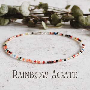 May include: A colourful rainbow agate beaded necklace with a silver clasp. The necklace is laid out on a white textured surface.