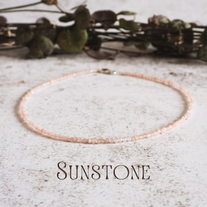 Peut inclure: Collier délicat en pierre de soleil. Le collier est composé d'un fil de petites perles facettées de couleur pêche. Le mot "SUNSTONE" est imprimé en marron, dans une police élégante, en dessous du collier. Le fond est une surface claire et texturée avec des feuilles séchées.