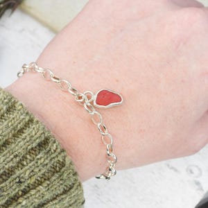 Könnte beinhalten: Silbernes Kettenarmband mit rotem Anhänger. Der Anhänger hat eine unregelmäßige Form, möglicherweise aus Meeresglas. Das Armband wird am Handgelenk getragen, mit einem grünen Pulloverärmel sichtbar.