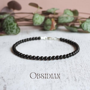 May include: A bracelet composed of small, round, polished black obsidian beads arranged in a single strand. The bracelet features a silver-toned metal clasp closure. The word OBSIDIAN is printed in a dark, serif font below the bracelet.