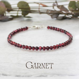 Puede incluir: Una delicada pulsera con pequeñas cuentas redondas de granate rojo oscuro, ensartadas. La pulsera tiene un cierre plateado. La palabra "GARNET" se muestra debajo en una fuente serif.