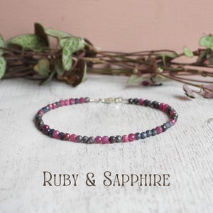 May include: A delicate beaded bracelet featuring alternating small, faceted ruby and sapphire gemstones. The bracelet is secured with a silver-toned metal lobster clasp. The text "RUBY & SAPPHIRE" is printed in a dark font below the jewellery piece.