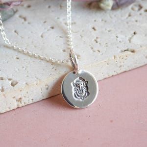 May include: A silver necklace featuring a small, circular pendant with a stamped Ganesha design. The pendant hangs from a delicate silver chain, showcasing a polished, reflective surface that highlights the intricate details of the embossed deity.
