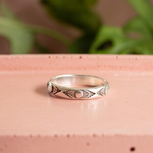 May include: A silver ring with an engraved pattern of suns and diamond shapes. The ring is set on a pink surface, with green foliage in the background. The ring is a band style, and the design is repeated around the entire ring.