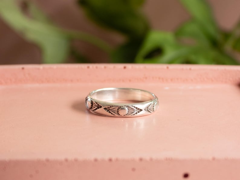 May include: A silver ring with an engraved pattern of suns and diamond shapes. The ring is set on a pink surface, with green foliage in the background. The ring is a band style, and the design is repeated around the entire ring.