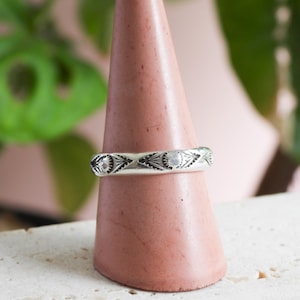 May include: A silver ring with an engraved geometric pattern. The ring is displayed on a pink cone-shaped stand. The design features repeating eye-like shapes and small circular details. The background includes green foliage and a pink surface.
