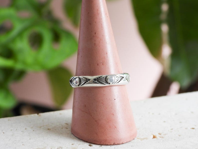 May include: A silver ring with an engraved geometric pattern. The ring is displayed on a pink cone-shaped stand. The design features repeating eye-like shapes and small circular details. The background includes green foliage and a pink surface.