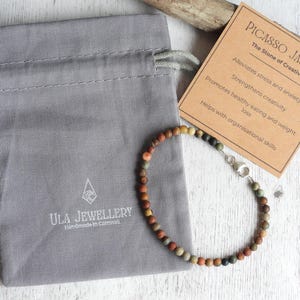 May include: A Picasso Jasper beaded bracelet with a silver clasp, presented with a gray drawstring bag and a card describing the stone's properties. The card reads "PICASSO JASPER The Stone of Creativity". The bag is branded "ULA JEWELLERY".