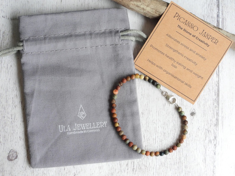 May include: A Picasso Jasper beaded bracelet with a silver clasp, presented with a gray drawstring bag and a card describing the stone's properties. The card reads "PICASSO JASPER The Stone of Creativity". The bag is branded "ULA JEWELLERY".