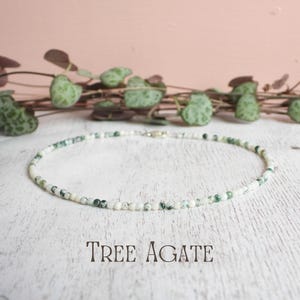 May include: A delicate necklace made of small, round, white and green beads. The necklace is displayed on a white wooden surface with the text "TREE AGATE" in a serif font. Green leafy vines are in the background.