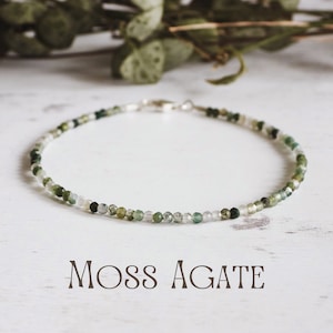 May include: A delicate bracelet made with moss agate beads. The beads are a mix of green and white, and the bracelet is strung on a silver chain. The bracelet is lying on a white wooden surface. Moss Agate.