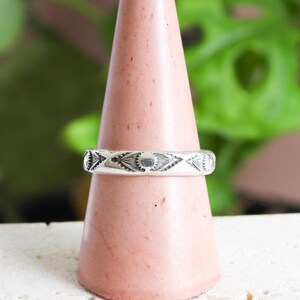 May include: A silver ring with an engraved geometric pattern, displayed on a pink cone-shaped stand. The ring's design includes repeating shapes and lines. The background features green foliage and a white surface.