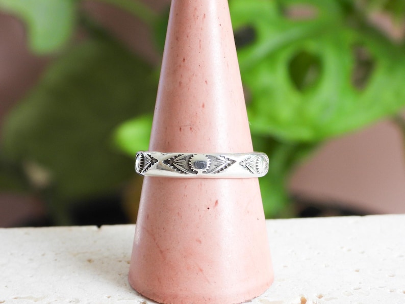 May include: A silver ring with an engraved geometric pattern, displayed on a pink cone-shaped stand. The ring's design includes repeating shapes and lines. The background features green foliage and a white surface.