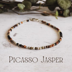 May include: A close-up of a necklace made with Picasso Jasper beads. The beads are a mix of brown, gray, white, and black. The necklace is on a white wooden surface. The text "PICASSO JASPER" is written below the necklace.