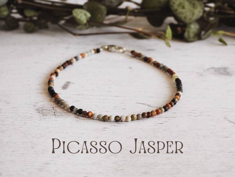 May include: A close-up of a necklace made with Picasso Jasper beads. The beads are a mix of brown, gray, white, and black. The necklace is on a white wooden surface. The text "PICASSO JASPER" is written below the necklace.